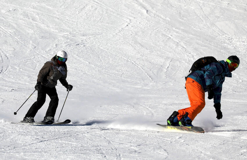Eine Frau auf Skier und ein Mann auf den Snowboard flitzen eine Skipiste hinunter.