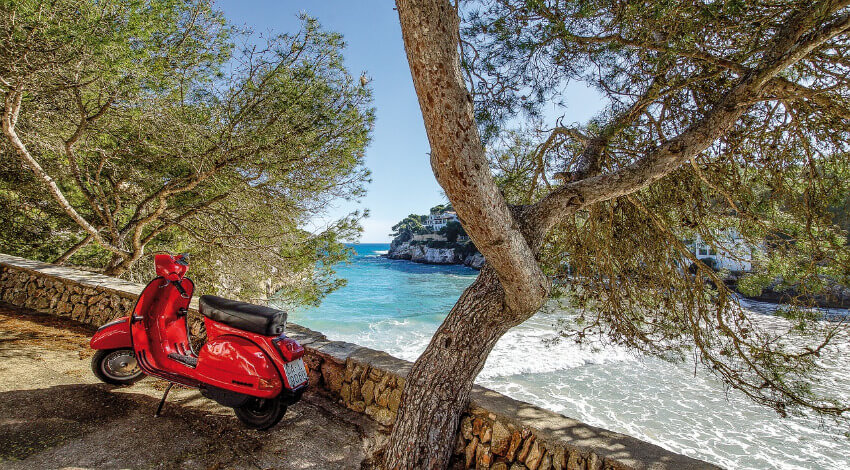 Ein roter Roller abgestellt am Straßenrand mit Blick auf die Bucht Cala Santanyí.